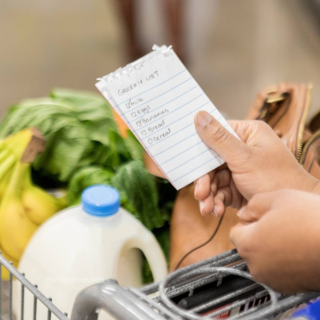 Hand holding a handwritten grocery list over a cart filled with milk, bananas, and fresh greens
