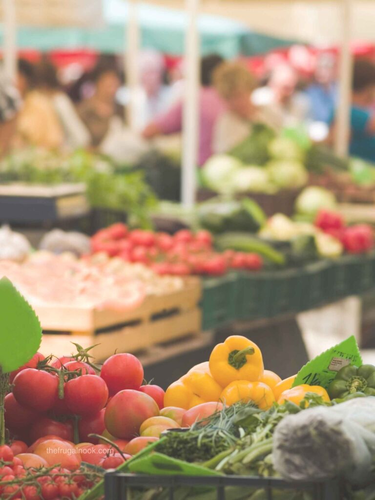 Farmers market stalls with tomatoes, yellow peppers, and fresh greens in the foreground and shoppers in the background