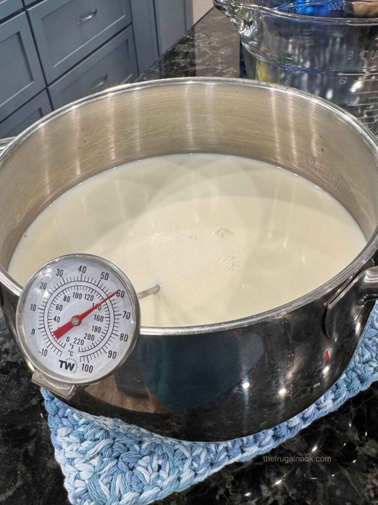 Goat milk heating in a stainless steel pot with a dial thermometer, set on a crocheted hot pad